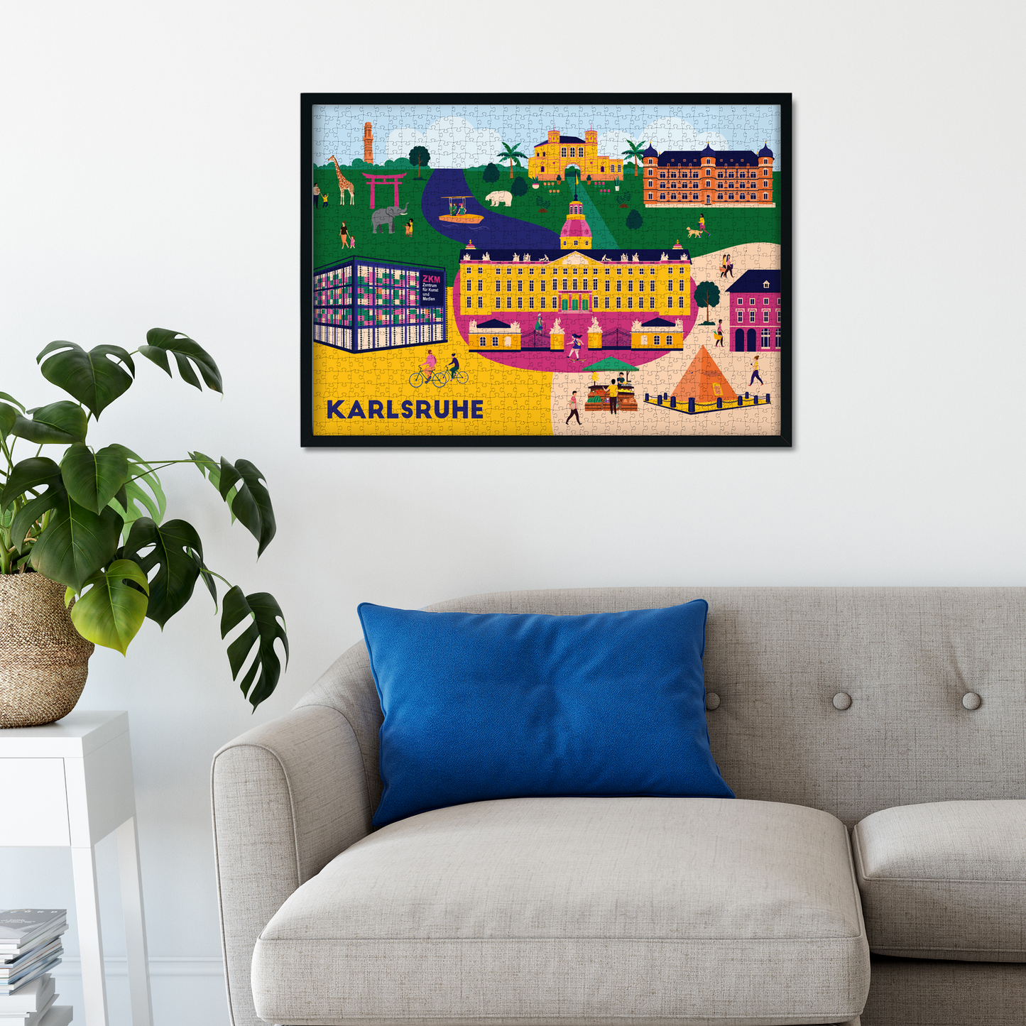 Gerahmtes Karlsruhe Puzzle mit farbenfroher Stadtillustration über einem modernen Sofa mit blauem Kissen – stilvoll dekoriert in einem minimalistischen Wohnzimmer.